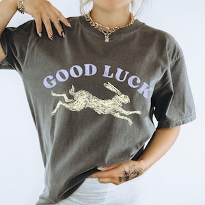 May include: Dark grey t-shirt featuring the words "GOOD LUCK" in light purple above a running rabbit illustration in yellow. The shirt has short sleeves and a crew neck.