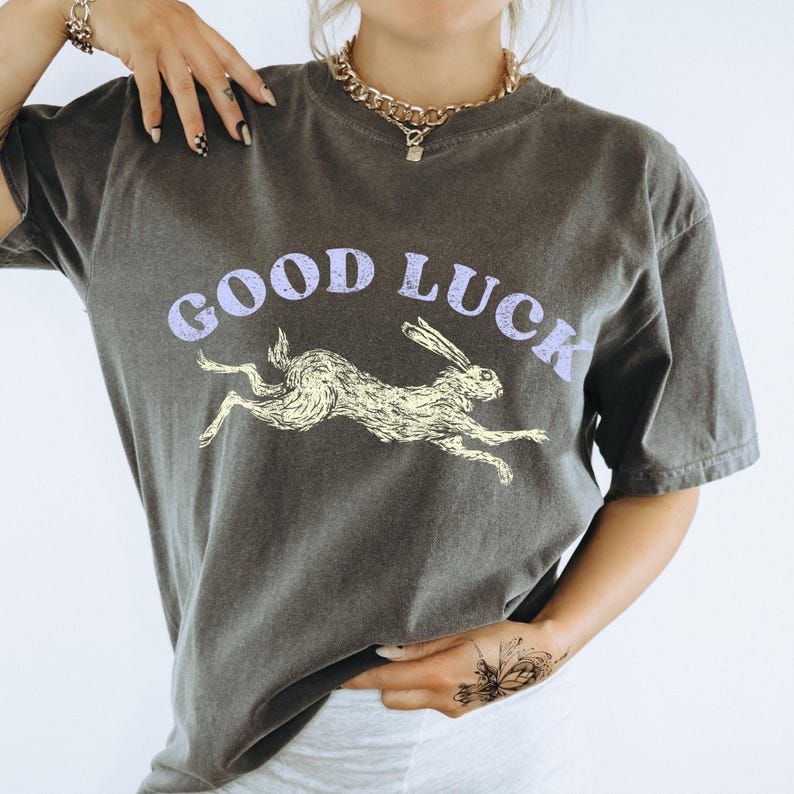 May include: Dark grey t-shirt featuring the words "GOOD LUCK" in light purple above a running rabbit illustration in yellow. The shirt has short sleeves and a crew neck.