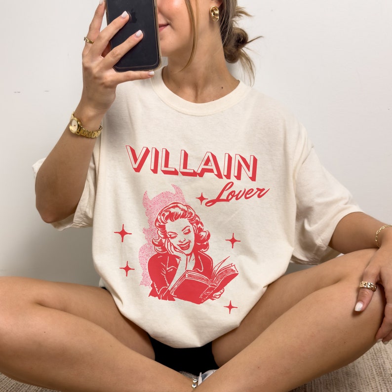 May include: A cream colored t-shirt with a red graphic print of a woman reading a book. The text "VILLAIN Lover" is printed in red above the graphic.