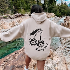 Op de afbeelding: Een cr&egrave;mekleurige hoodie met een zwarte afbeelding van twee kersen met het nummer 8 op elk. De tekst "Lucky you!" staat onder de kersen.