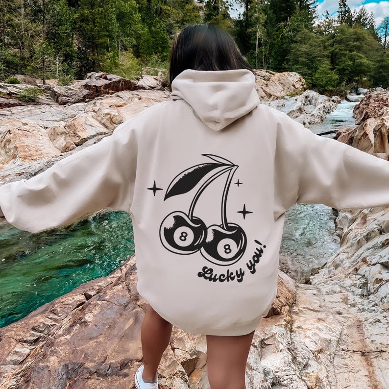 Op de afbeelding: Een cr&egrave;mekleurige hoodie met een zwarte afbeelding van twee kersen met het nummer 8 op elk. De tekst "Lucky you!" staat onder de kersen.
