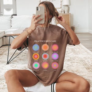 May include: A brown t-shirt with the text "PRACTICE SELF-CARE" and a graphic of nine colorful circles with text describing self-care activities, such as "read a book", "drink water", "go on a walk", "journal", "take a nap", "listen to music", "enjoy the sun", "meditate", and "smile".