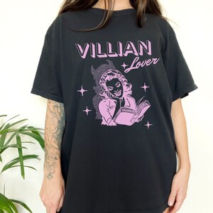 May include: Black t-shirt with a pink graphic of a woman reading a book. The text "VILLIAN Lover" is printed in pink above the graphic.