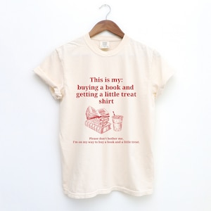 May include: A beige t-shirt with a red graphic of a stack of books and a cup of coffee. The text on the shirt reads "This is my: buying a book and getting a little treat shirt. Please don't bother me, I'm on my way to buy a book and a little treat."