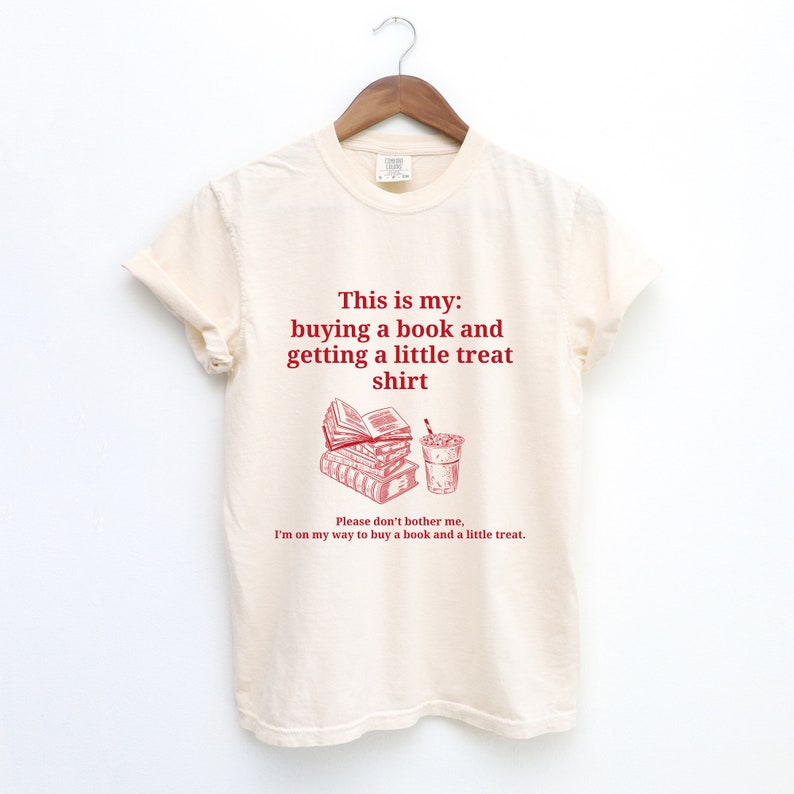 May include: A beige t-shirt with a red graphic of a stack of books and a cup of coffee. The text on the shirt reads "This is my: buying a book and getting a little treat shirt. Please don't bother me, I'm on my way to buy a book and a little treat."