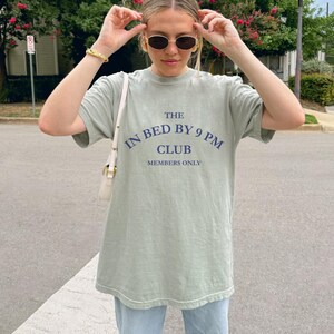 May include: Sage green t-shirt with the text "THE IN BED BY 9 PM CLUB MEMBERS ONLY" in navy blue. The shirt is a relaxed fit and appears to be made of a soft, comfortable material. The shirt is being worn outdoors.