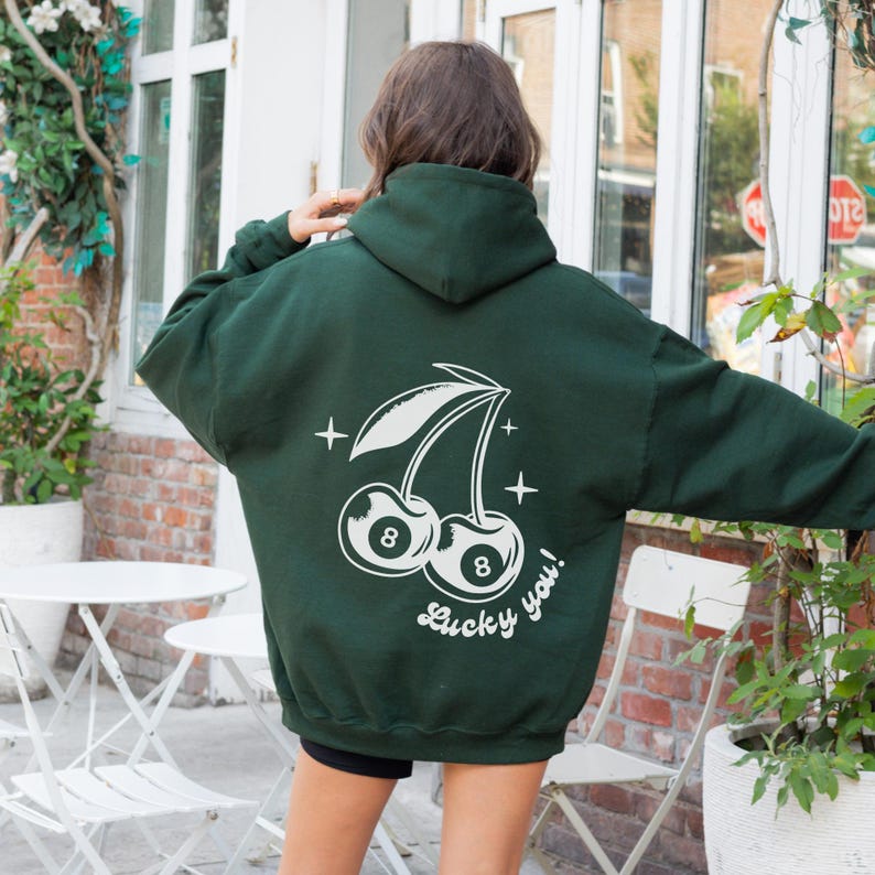 Op de afbeelding: Een donkergroen hoodie met een witte afbeelding van twee kersen met het nummer 8 erop. De tekst "Lucky you!" staat onder de kersen.