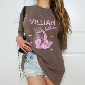 May include: A brown oversized t-shirt with a pink graphic of a woman reading a book. The text on the shirt reads "Villian Lover".