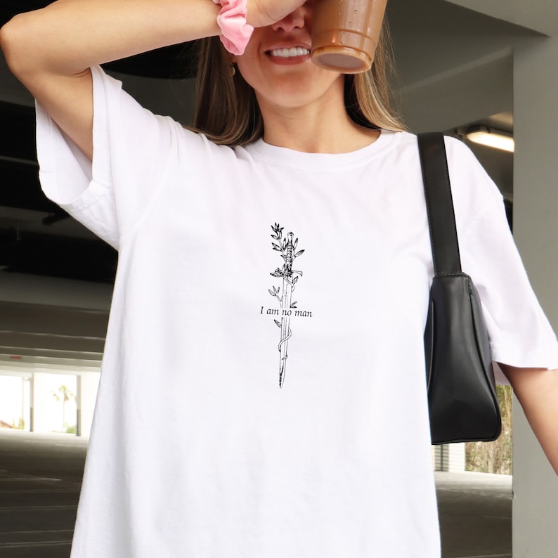 Pode incluir: Camiseta branca com um gr&aacute;fico preto de uma espada com videiras e o texto "I am no man".