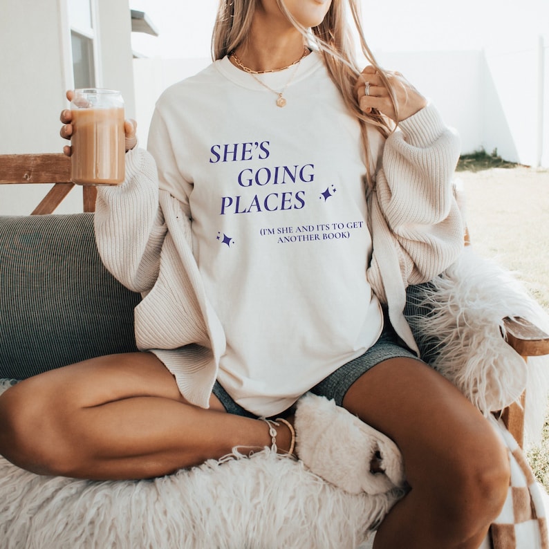 Puede incluir: Sudadera blanca con el texto "She's Going Places (I'm She and It's to Get Another Book)" en letras moradas.