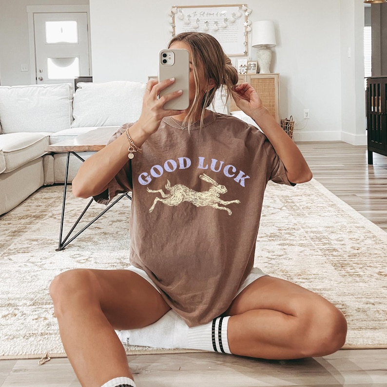 May include: A brown t-shirt featuring the words "GOOD LUCK" in lavender above a gold rabbit graphic. The shirt is paired with white shorts and black and white striped socks. The person is holding a phone.