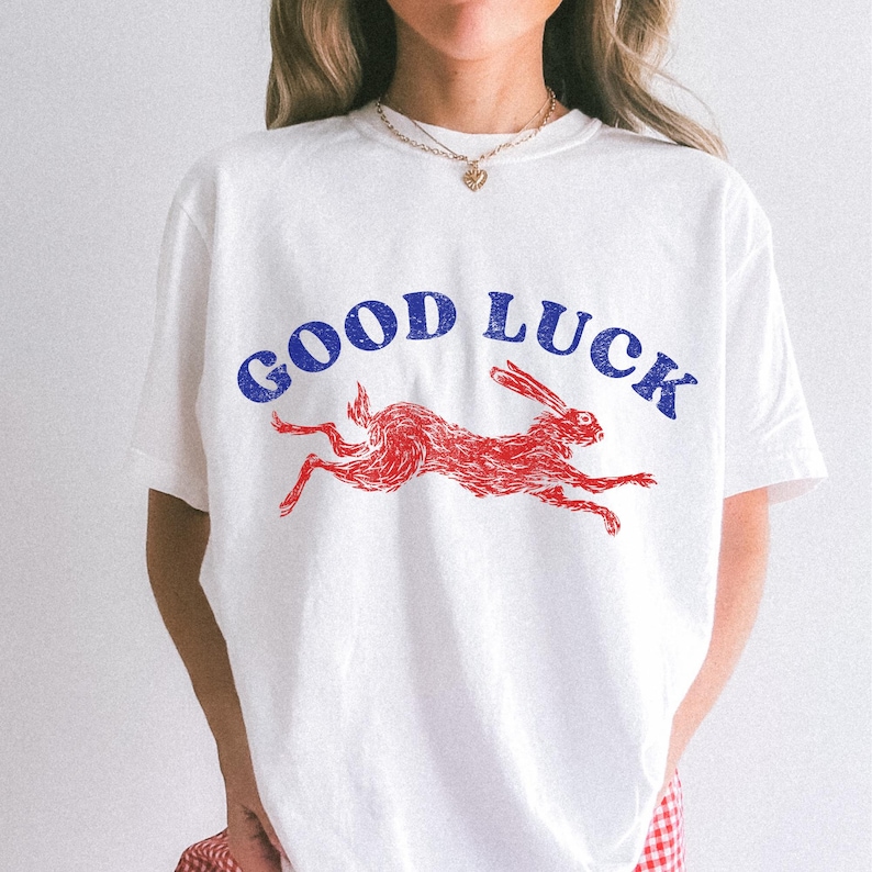 May include: White t-shirt featuring a red rabbit graphic and the blue text "GOOD LUCK". The rabbit is in motion. The shirt is a casual style.