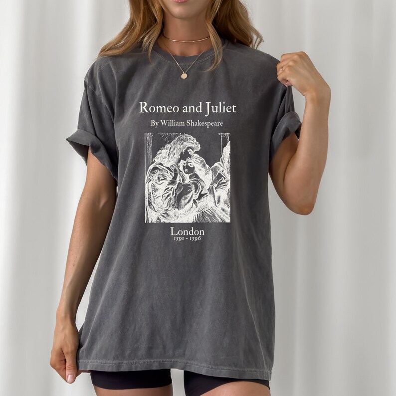May include: A dark gray t-shirt with a white graphic print of a book cover. The book cover features a black and white illustration of two people embracing. The title of the book is "Romeo and Juliet" by William Shakespeare. The text "London 1591 - 1596" is printed below the illustration.