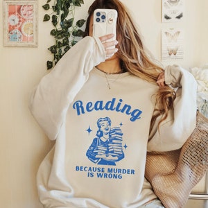 May include: A light beige sweatshirt with the text "Reading because murder is wrong" in blue. A retro illustration of a woman holding a stack of books is above the text.