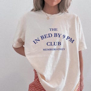 May include: Cream-colored t-shirt with the text "THE IN BED BY 9 PM CLUB MEMBERS ONLY" in navy blue. The shirt has a crew neck and short sleeves. The text is arched above the word "CLUB".