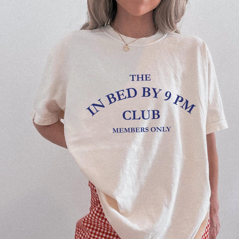 May include: Cream-colored t-shirt with the text "THE IN BED BY 9 PM CLUB MEMBERS ONLY" in navy blue. The shirt has a crew neck and short sleeves. The text is arched above the word "CLUB".