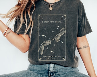 May include: A dark gray t-shirt with a vintage-style graphic featuring two hands reaching towards each other with stars and moons in the background. The text "I defy you, stars." is printed above the graphic, and "Romeo and Juliet Act 5, Scene I" is printed below.