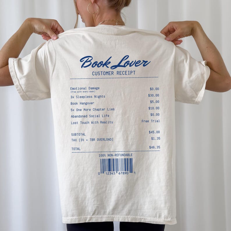 May include: Cream-colored t-shirt with the text "Book Lover" and a customer receipt design printed in blue. The receipt lists humorous "charges" like "Emotional Damage" and "3x Sleepless Nights."