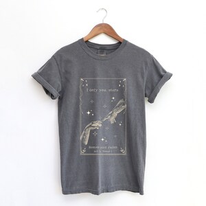 May include: A dark grey t-shirt featuring a graphic print of two hands reaching towards each other with stars in the background. The text "I defy you, stars" and "Romeo and Juliet, Act 3, Scene 1" is printed below the graphic.