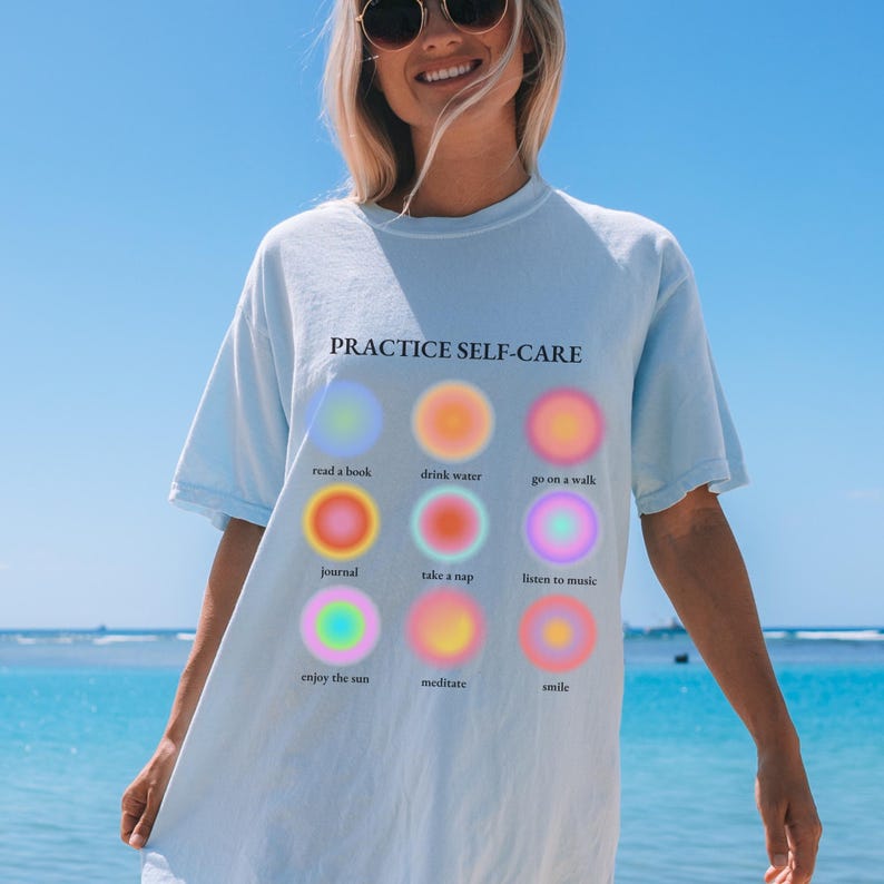 May include: A light blue t-shirt with the text "PRACTICE SELF-CARE" printed on the front. The shirt features nine colorful circles with text inside each circle, including "read a book", "drink water", "go on a walk", "journal", "take a nap", "listen to music", "enjoy the sun", "meditate", and "smile".