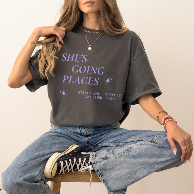 Puede incluir: Una camiseta gris antracita con el texto "She's Going Places (I'm She and It's to Get Another Book)" en letras moradas. La camiseta tiene un corte casual y mangas cortas.