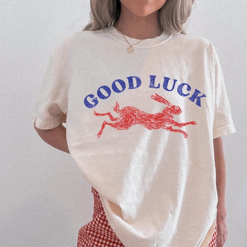 May include: Cream-coloured t-shirt featuring the words "GOOD LUCK" in blue above a red illustration of a running rabbit. The shirt is a casual style, suitable for everyday wear.