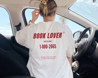 May include: Cream-coloured t-shirt with the words "BOOK LOVER" and "HOTLINE 1-800-2665" printed in red. The text "THE ONLY THING YOU NEED IN LIFE IS A GOOD BOOK" is printed below. The shirt is being worn in a car.
