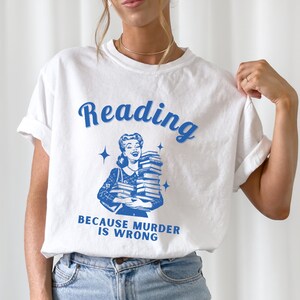 May include: White t-shirt with a blue graphic of a woman holding a stack of books. The text "Reading because murder is wrong" is printed below the graphic.