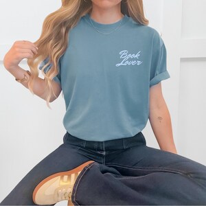 May include: A blue-green t-shirt with the words "Book Lover" in a cursive font. The shirt has rolled-up sleeves and is paired with dark blue jeans. The person is wearing a gold bracelet and a necklace.