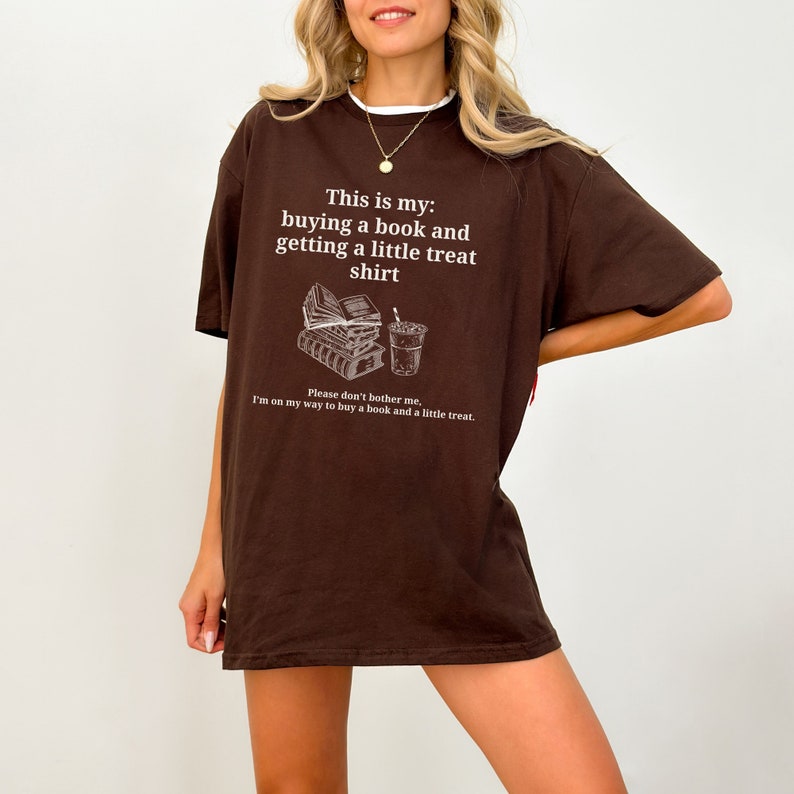 May include: Brown oversized t-shirt with a graphic of a stack of books and a cup of coffee. The text on the shirt reads "This is my: buying a book and getting a little treat shirt. Please don't bother me, I'm on my way to buy a book and a little treat."