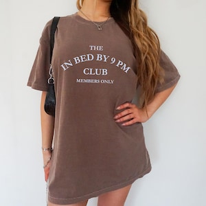 May include: Brown t-shirt with the text "THE IN BED BY 9 PM CLUB MEMBERS ONLY" in white. The shirt has short sleeves and a relaxed fit. The model is wearing a black purse.