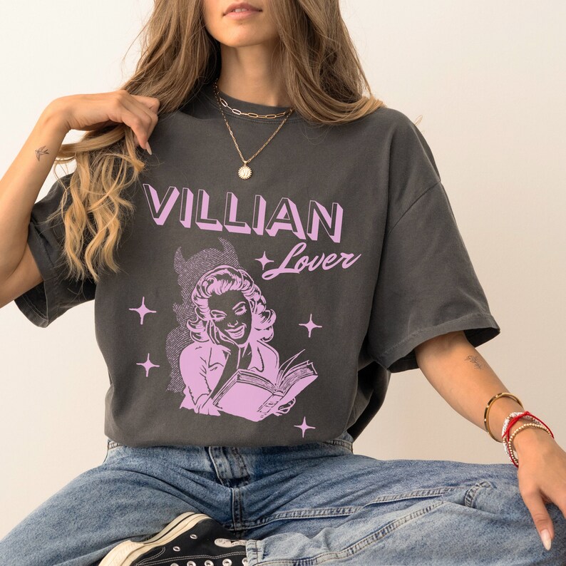 May include: A charcoal grey t-shirt with a pink graphic of a woman reading a book. The text "VILLIAN Lover" is printed on the shirt.