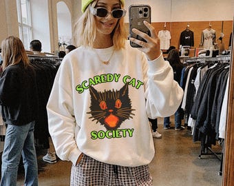 Könnte beinhalten: Weißes Sweatshirt mit einem schwarzen Katzen-Grafikdesign und dem grünen Schriftzug "Scaredy Cat Society". Die Katze hat orangefarbene Augen und einen Mund. Die Person trägt das Sweatshirt in einem Geschäft.
