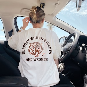 May include: Cream-colored t-shirt with a brown tiger graphic and the text "SUPPORT WOMEN'S RIGHTS AND WRONGS" in a semi-circular design. The tiger graphic is roaring, and the shirt is worn in a car.