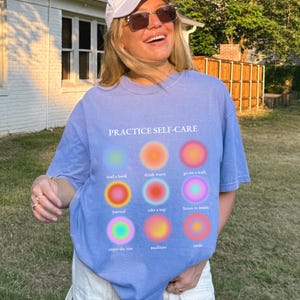 May include: A light blue t-shirt with the text "PRACTICE SELF-CARE" printed on the front. The shirt features nine colorful circles with text inside each circle, including "read a book", "drink water", "go on a walk", "journal", "take a nap", "listen to music", "enjoy the sun", "meditate", and "smile".