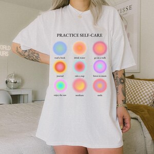 May include: White t-shirt with the text "PRACTICE SELF-CARE" printed on the front. The text is surrounded by nine colorful circles, each with a different self-care activity listed below it: read a book, drink water, go on a walk, journal, take a nap, listen to music, enjoy the sun, meditate, smile.