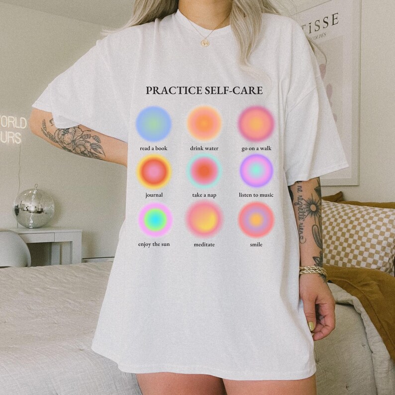 May include: White t-shirt with the text "PRACTICE SELF-CARE" printed on the front. The text is surrounded by nine colorful circles, each with a different self-care activity listed below it: read a book, drink water, go on a walk, journal, take a nap, listen to music, enjoy the sun, meditate, smile.