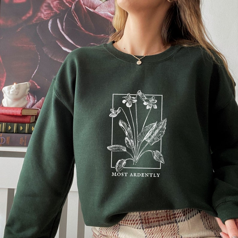 May include: A dark green sweatshirt with a white floral illustration and the text "MOST ARDENTLY" printed on the front.