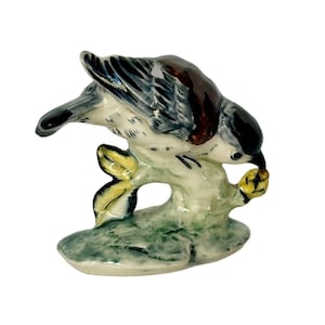 May include: A ceramic figurine of a blue and gray bird perched on a branch, with a brown head and yellow accents. The bird is depicted eating a yellow berry.