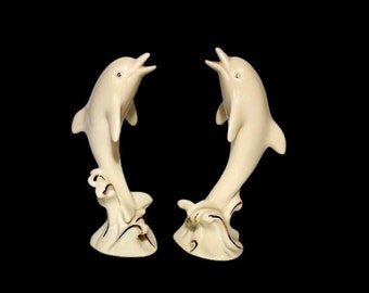 May include: Two white porcelain dolphin figurines leaping out of the water. The dolphins are facing each other and have gold accents.