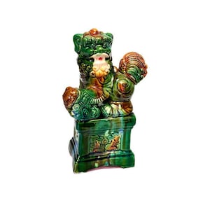 May include: A green and brown ceramic figurine of a mythical lion-like creature with a ball in its paw, sitting on a green and brown base.