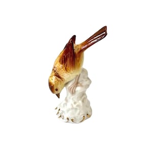 May include: A ceramic bird figurine with a downward gaze, perched on a white, textured base. The bird features a blend of brown and tan hues, with a detailed tail and wings. The base is adorned with gold accents, creating a decorative piece.
