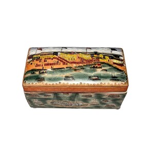 May include: Rectangular decorative box with a detailed landscape scene. The lid features a waterfront with buildings, flags, and boats. The box's sides have a mirrored image of boats. The color palette includes orange, yellow, green, and brown.