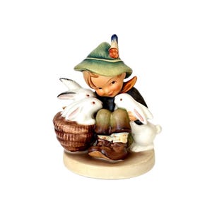 May include: A ceramic figurine of a young person in a green hat and dark coat, surrounded by three white rabbits. One rabbit is in a brown basket. The figurine has a light brown base.