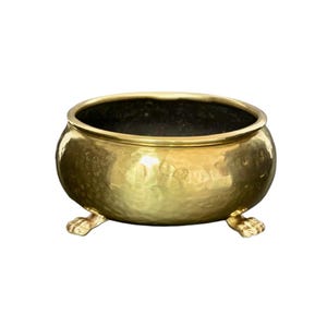 May include: A gold hammered brass planter with three decorative feet. The planter is round and has a smooth, shiny finish.