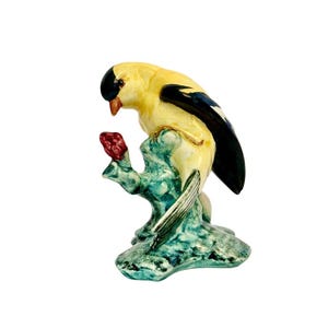 May include: A ceramic figurine of a yellow bird with black markings perched on a green branch with a red flower. The bird is depicted in a lifelike pose, with its head tilted and its beak open.