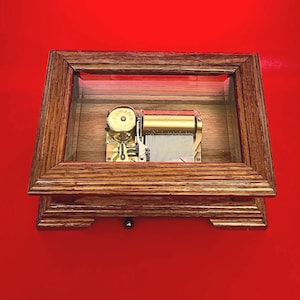 May include: A vintage music box with a wooden frame and a glass top. The music box mechanism is visible inside, featuring gold-colored components and a cylinder. The word "Romance" is printed on the top. The frame is made of dark wood.