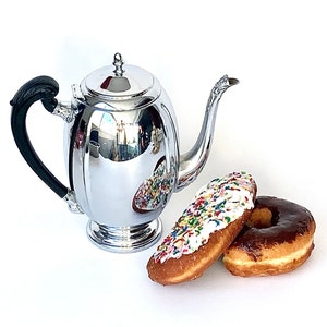 May include: A silver coffee pot with a black handle and a shiny finish. The pot is sitting on a white surface with two donuts, one with white frosting and sprinkles and the other with chocolate frosting.