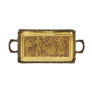 May include: A rectangular gold-toned metal tray with ornate handles and a detailed border. The tray's surface features an engraved scene of Egyptian figures and hieroglyphs. The handles have a decorative, openwork design.