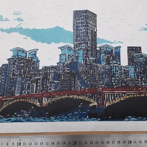 Peut inclure: Une estampe linogravure représentant une ville avec un pont et une rivière. Les bâtiments sont un mélange de bleu, gris et noir, et le pont est rouge. Le ciel est bleu avec des nuages blancs.